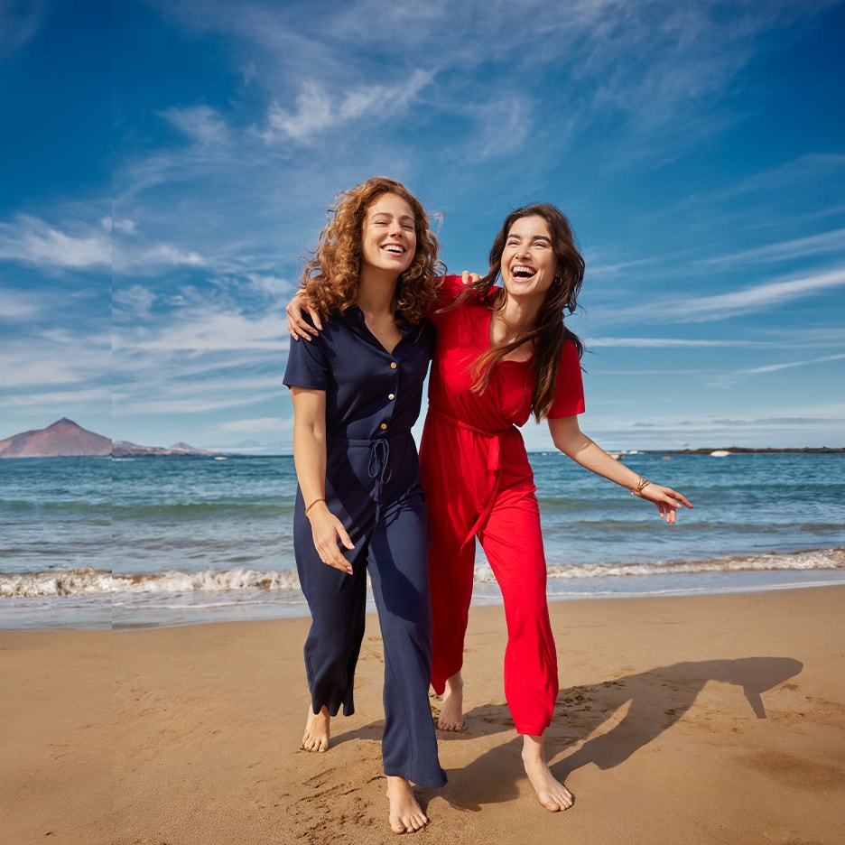 Zwei Frauen in Sommer-Overalls, einer blau und einer rot, gehen lachend am Strand entlang.