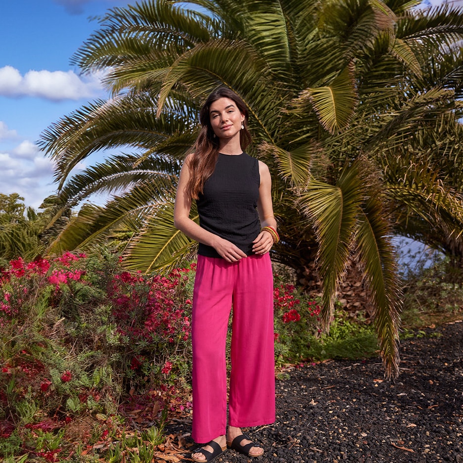 Frau in schwarzem Top und fuchsiafarbener Hose, mit Palmen und roten Blumen im Hintergrund.