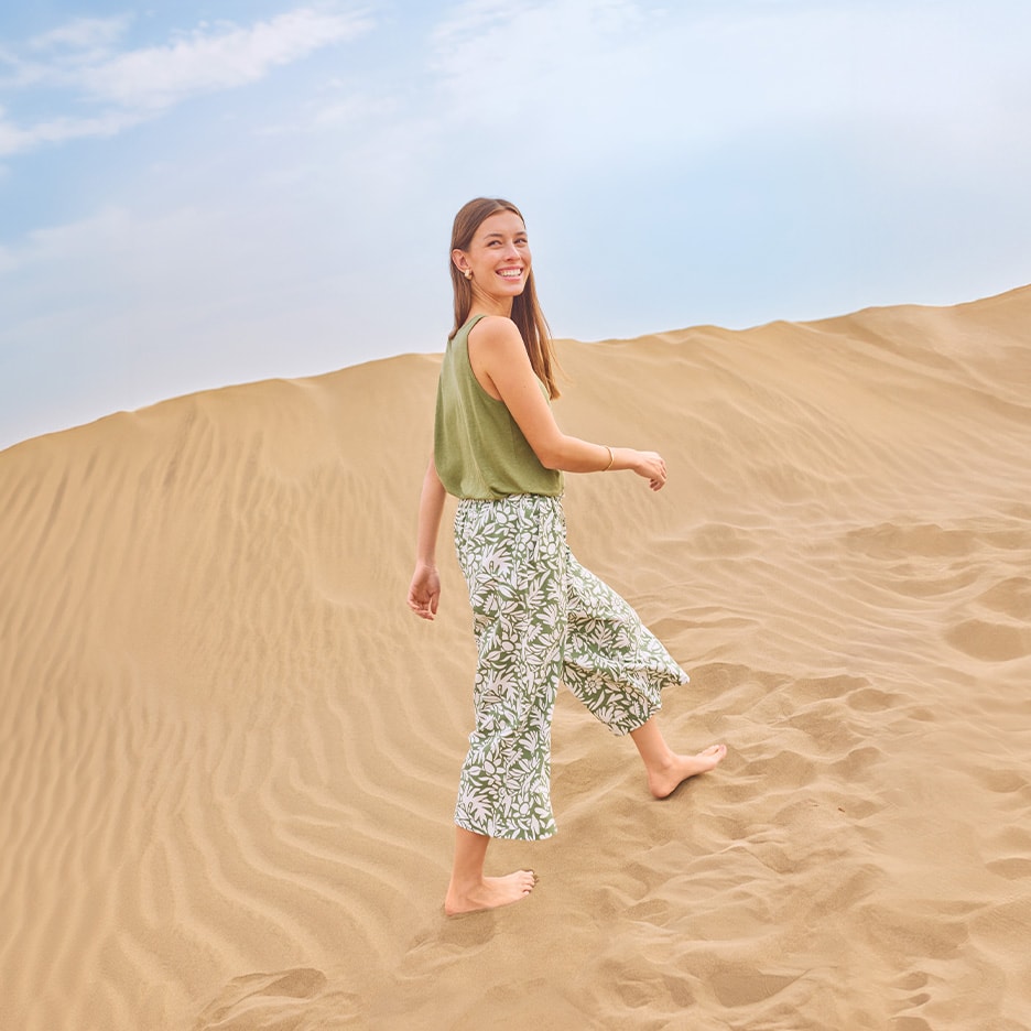 Lächelnde Frau in grünem Top und floral gemusterter Hose, die eine Sanddüne hinaufgeht.