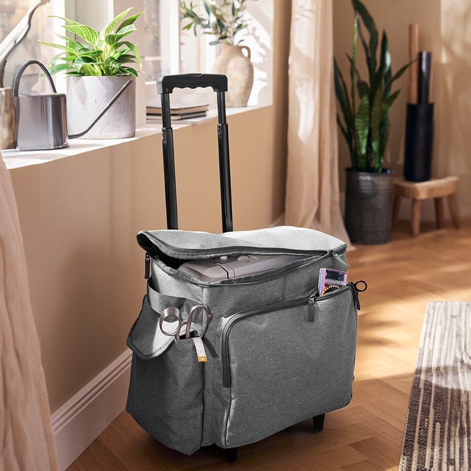 Graue Nähmaschinen-Trolleytasche mit Werkzeug, neben einem Fenster mit Pflanzen.