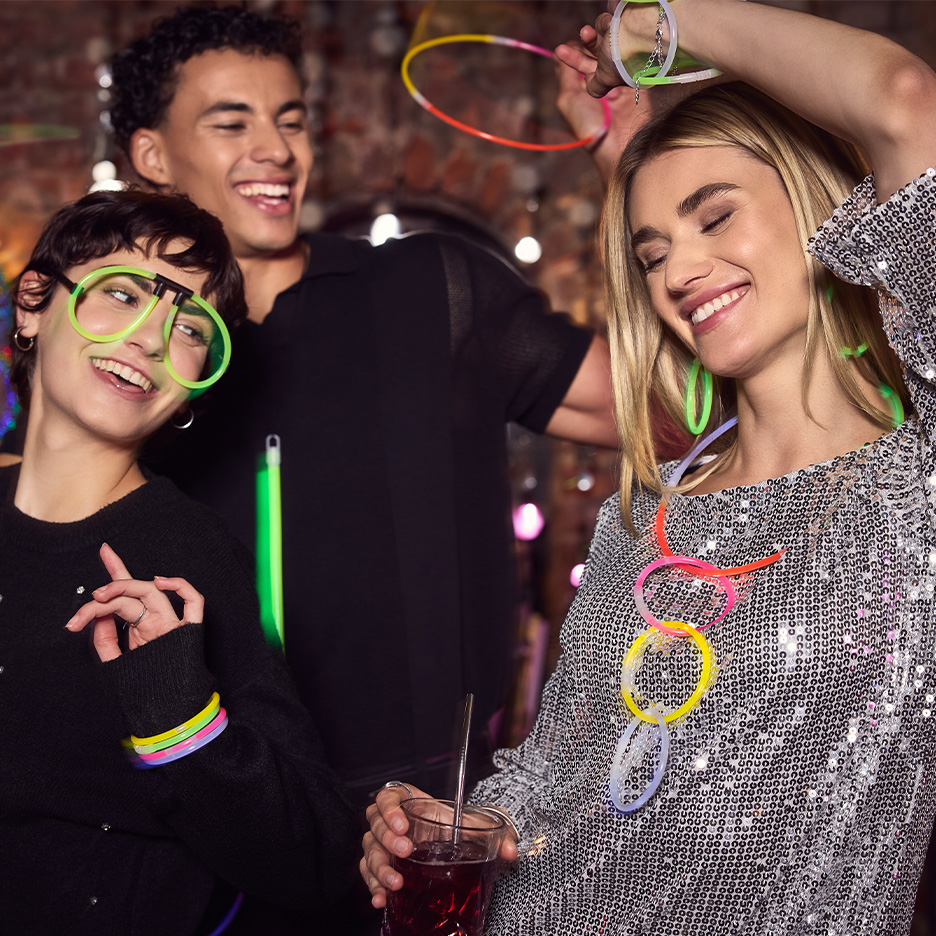 Drei lächelnde Freunde auf einer Party, die leuchtende Brillen und Schmuck tragen.