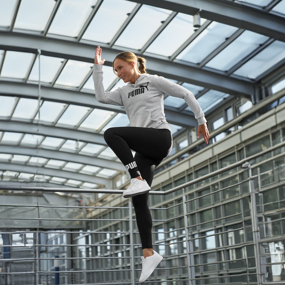 Frau in grauem Kapuzenpullover und schwarzen Leggings springt in einem modernen Gebäude.