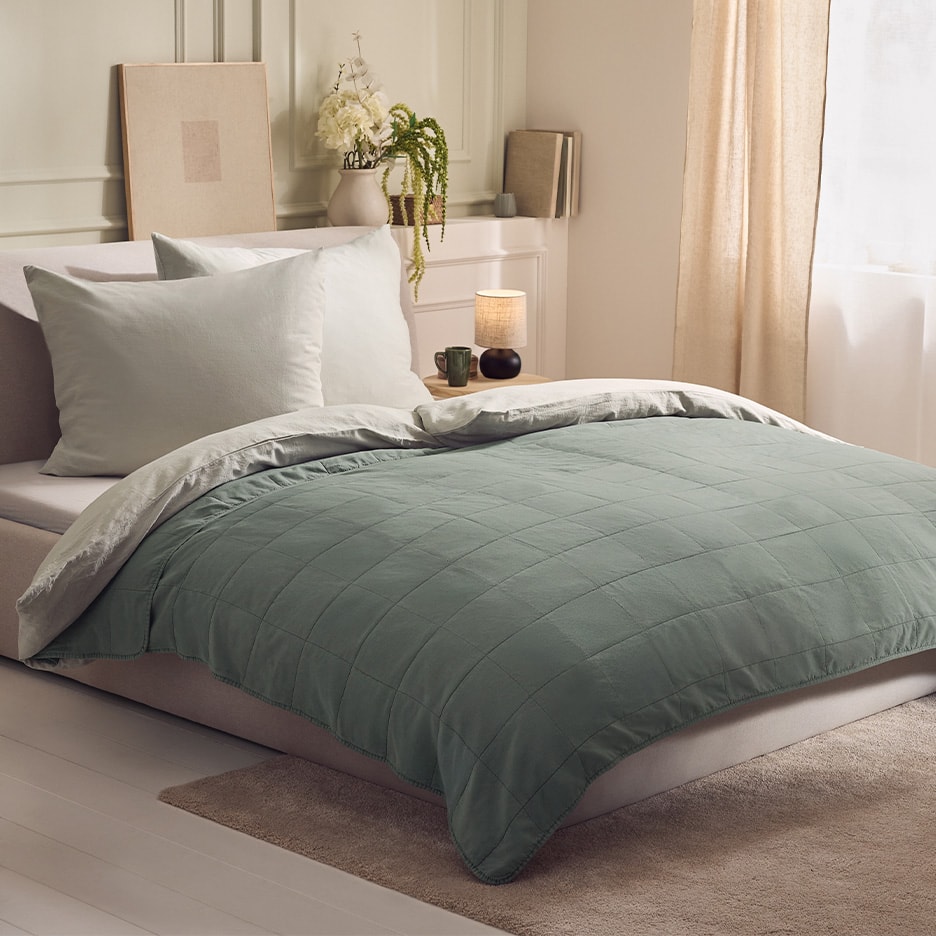 Bett mit gesteppter grüner Bettdecke und weißen Kissen in einem gemütlichen Schlafzimmer.