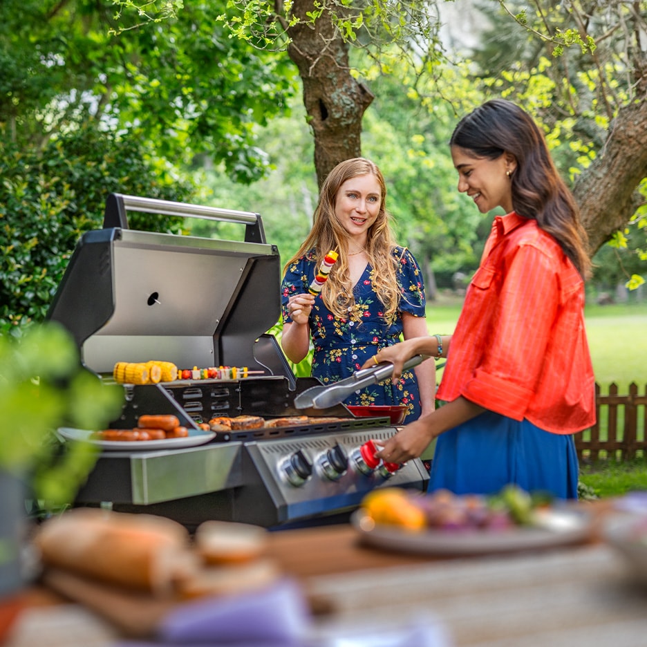 Zwei Frauen grillen im Garten, mit Spießen und Würstchen auf dem Grill.