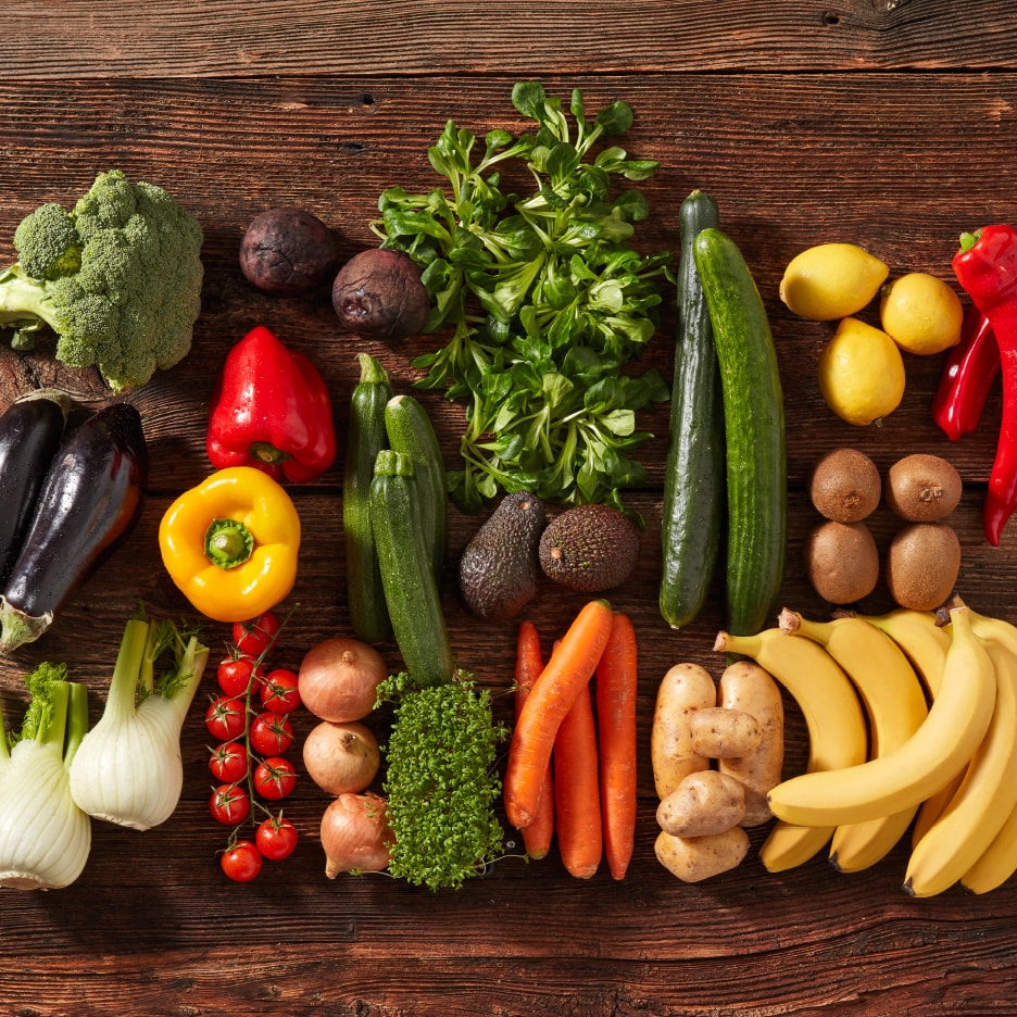 Eine Vielfalt an frischem Obst und Gemüse, darunter Brokkoli, Auberginen, Paprika, Zucchini, Karotten und Bananen, auf einem Holztisch arrangiert.