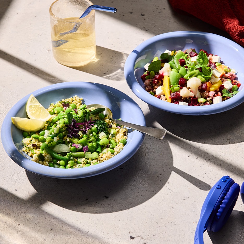 Zwei bunte Getreideschalen mit Gemüse und ein Glas Wasser auf einer sonnigen Oberfläche.