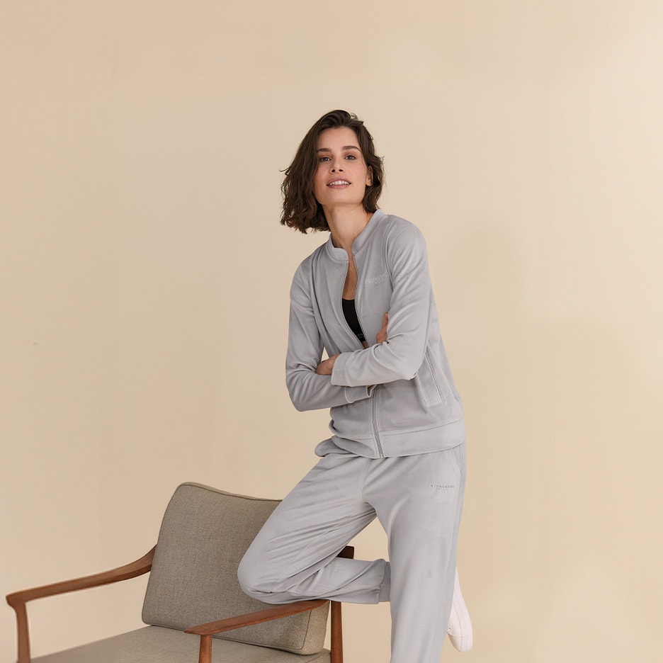 Frau in hellgrauem Trainingsanzug, Reißverschlussjacke und Hose, posiert mit einem Sessel.