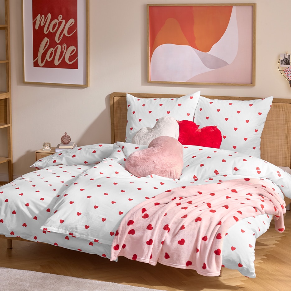 Bettwäsche mit roten Herzen, dazu passende Kissen und eine Decke in einem Schlafzimmer mit romantischer Dekoration.