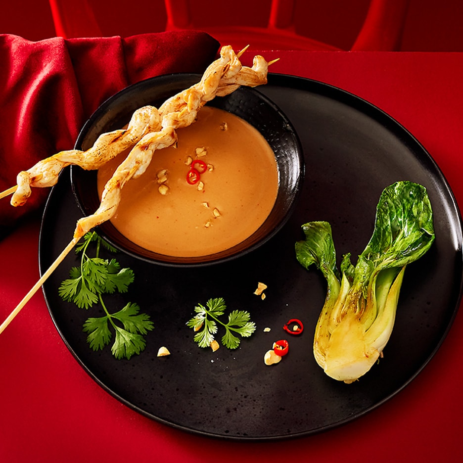 Hähnchenspieße mit Satay-Sauce, Pak Choi und Koriander auf einem schwarzen Teller.