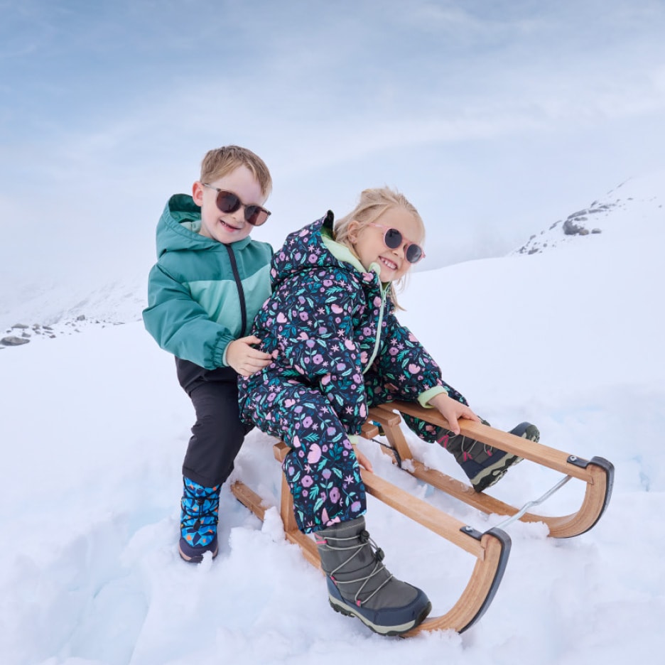 Zwei lächelnde Kinder in Schneeanzügen und Sonnenbrillen auf einem Schlitten.