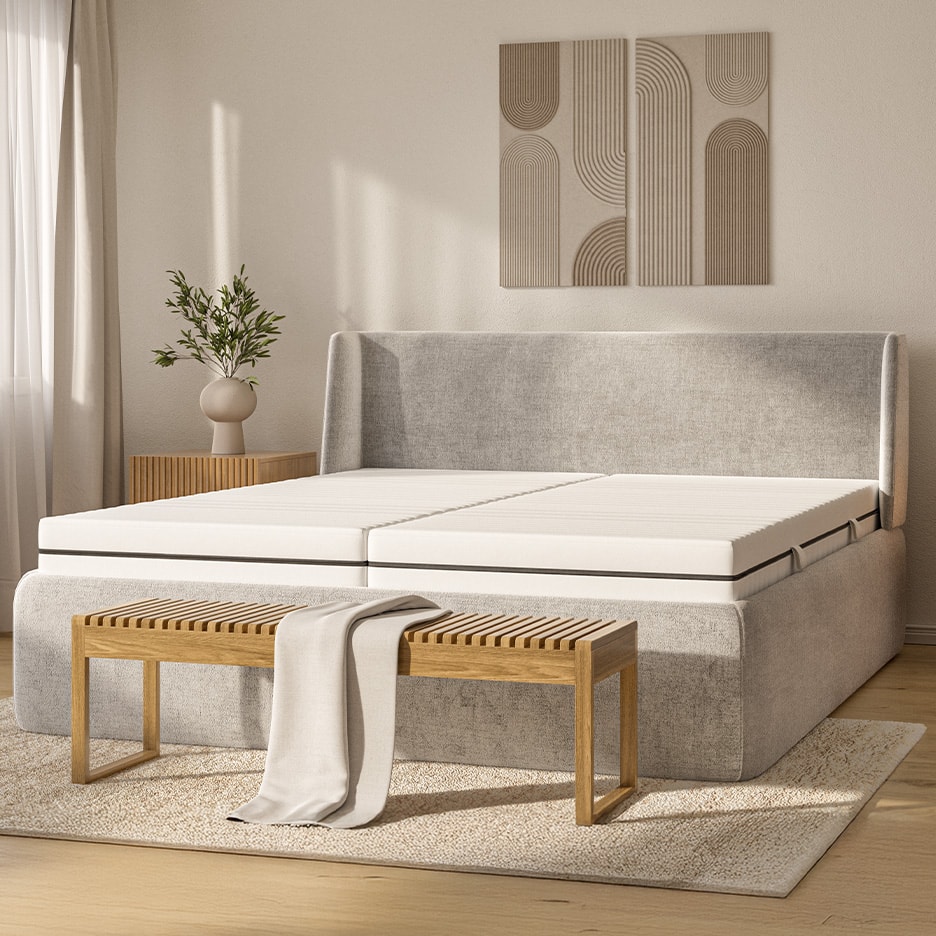 Graues Polsterbett mit weißen Matratzen, Holzbank und abstrakten Wanddekorationen.