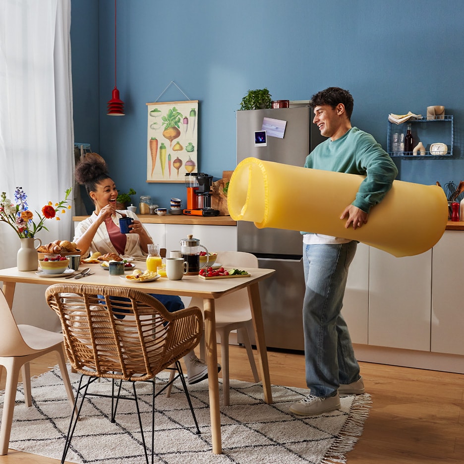 Ein Paar beim Frühstück, der Mann trägt eine riesige Nudelrolle.