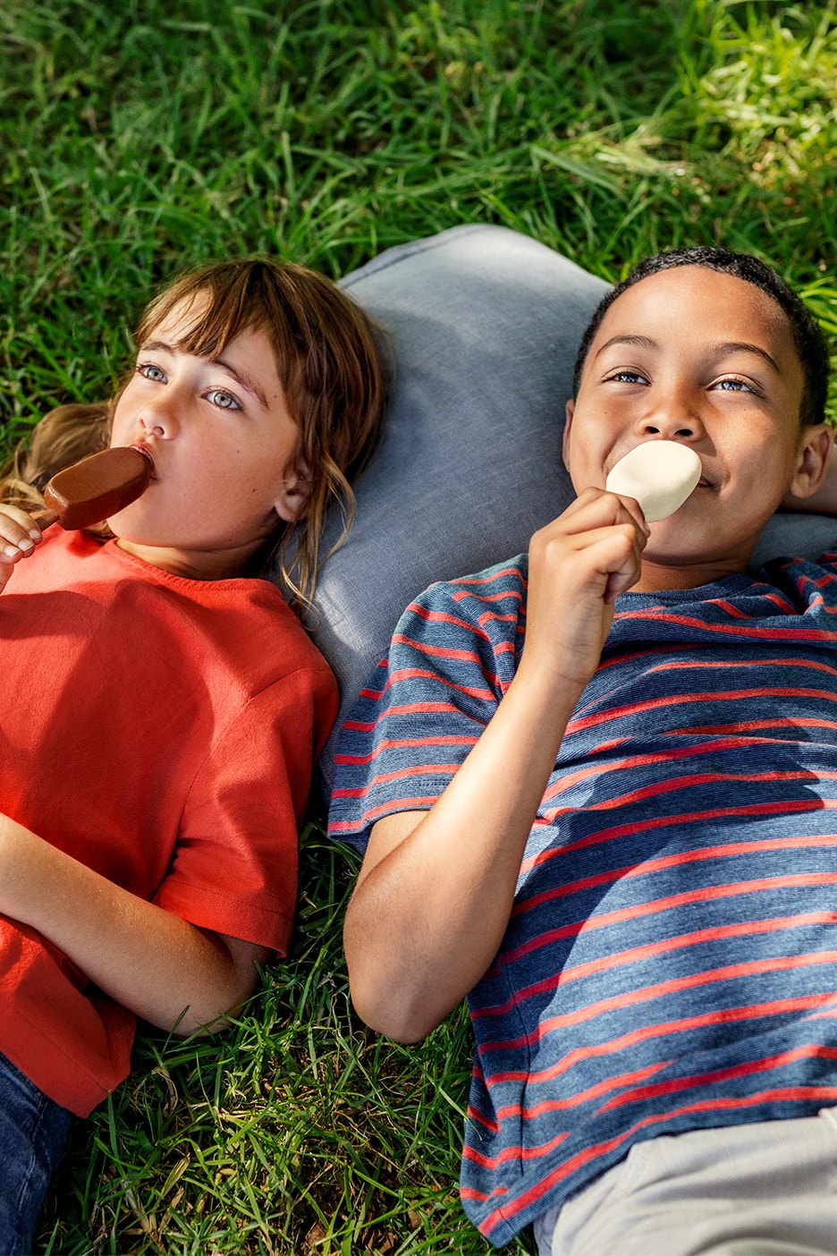 Zwei Kinder liegen im Gras und essen Eis am Stiel.