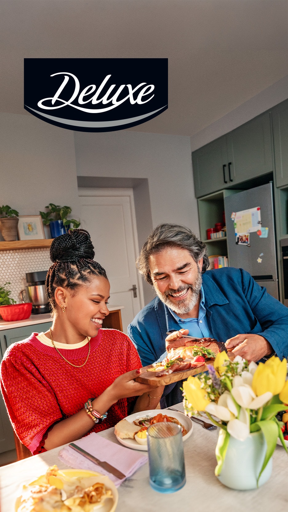 Un couple souriant partage un plateau de charcuterie et de fromages, avec des viennoiseries et des fruits sur la table.