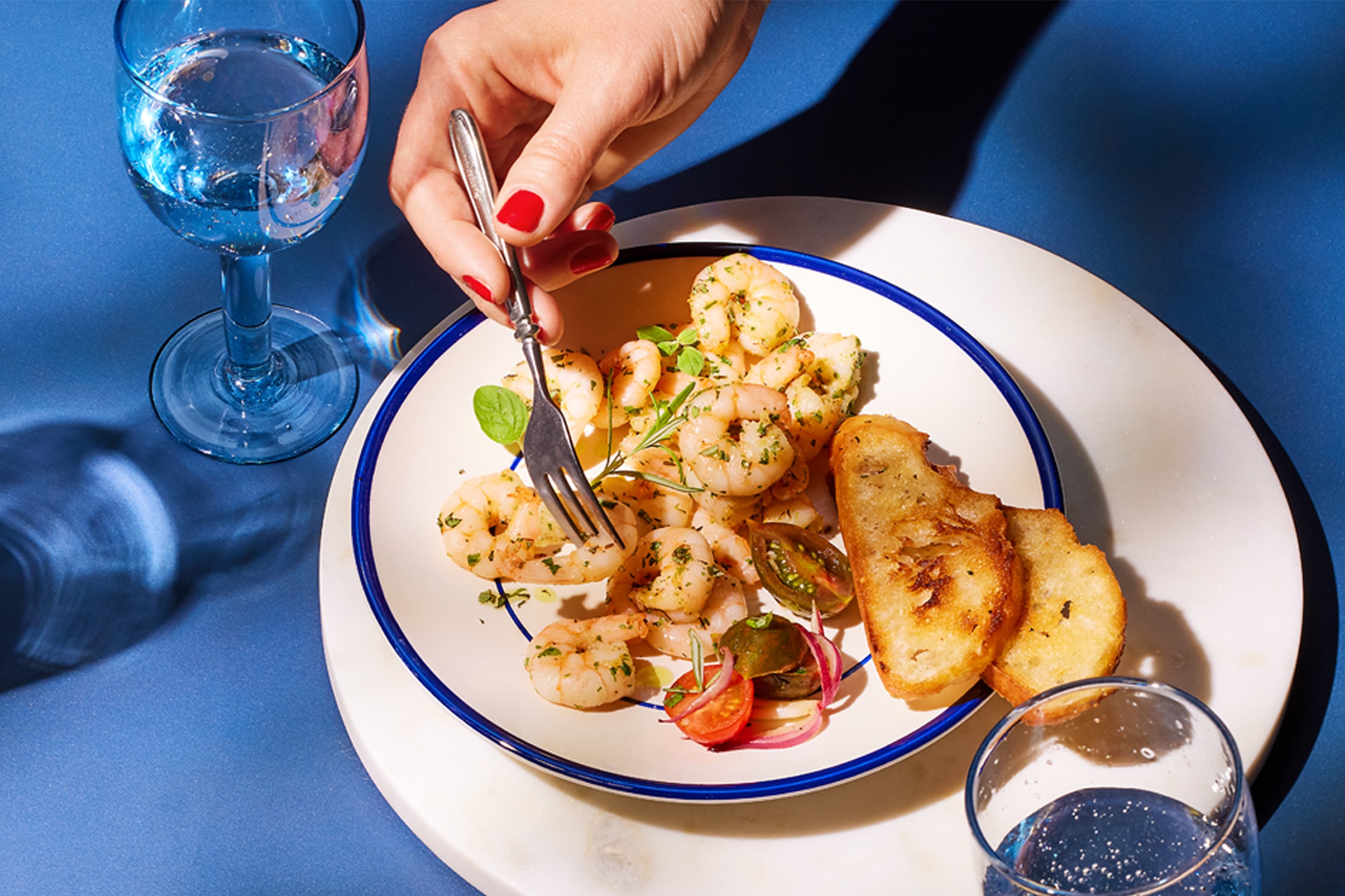 Gebratene Garnelen mit Kräutern, Kirschtomaten, roten Zwiebeln und geröstetem Brot, dazu ein Glas Wasser.