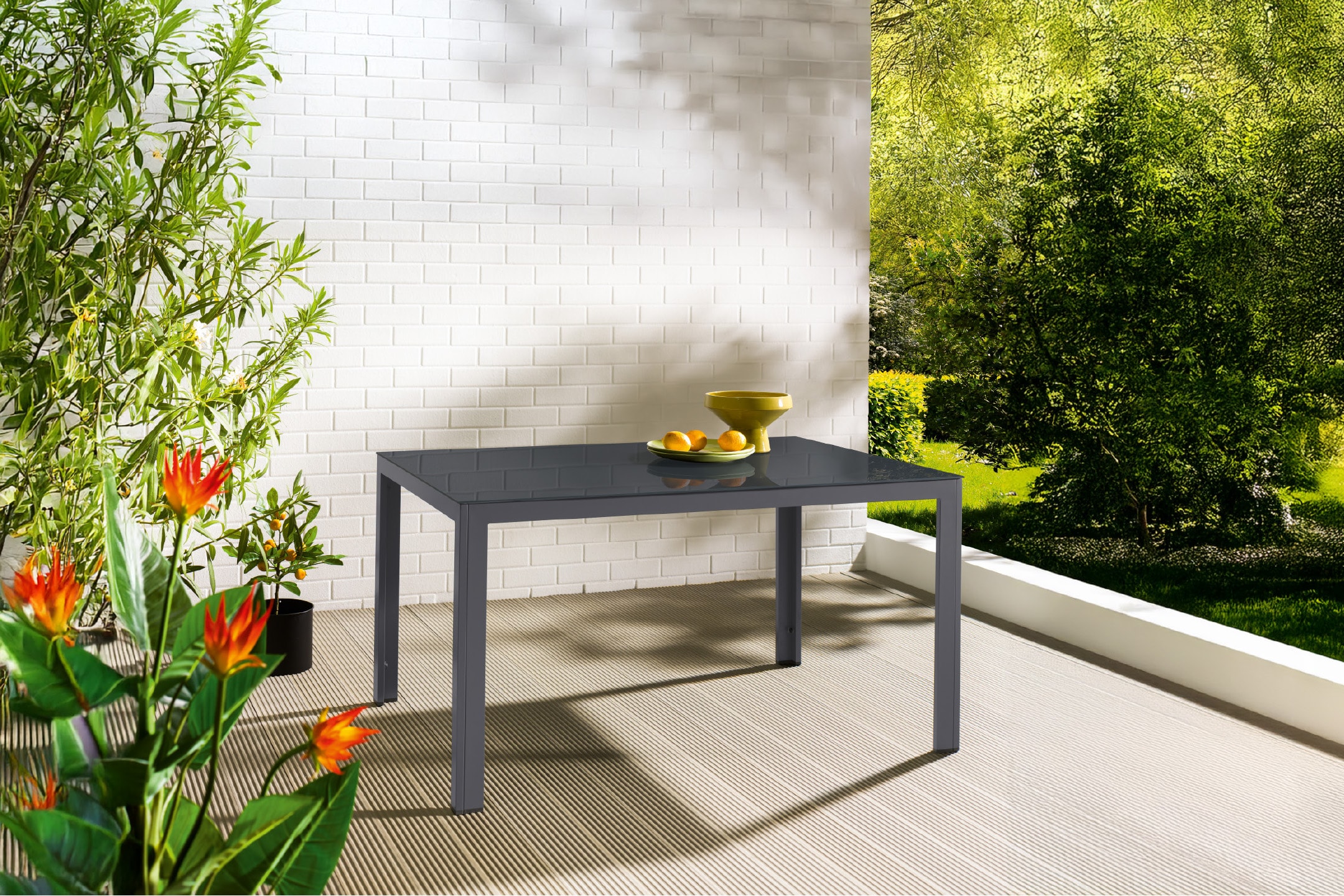 Tavolo da esterno grigio con piano in vetro su un balcone con piante e alberi.