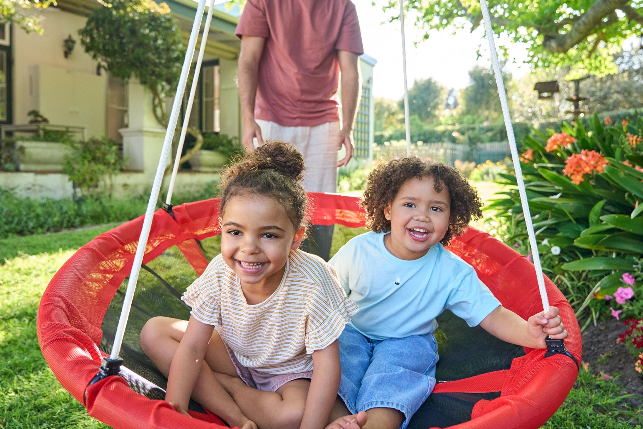 Zwei lächelnde Kinder auf einer roten Rundschaukel im Garten, mit einem Erwachsenen im Hintergrund.