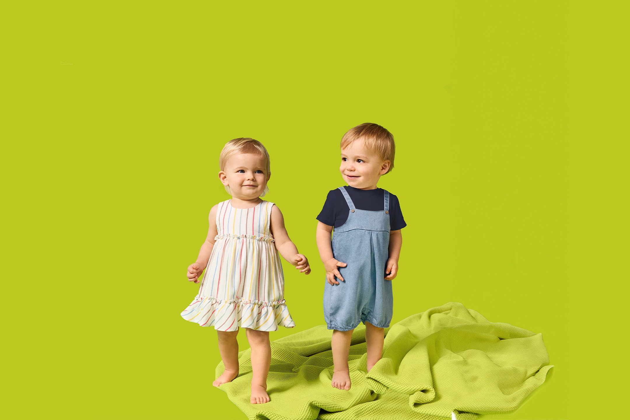 Zwei Kleinkinder, ein Mädchen im gestreiften Kleid und ein Junge im Jeans-Overall, stehen auf einer grünen Decke.