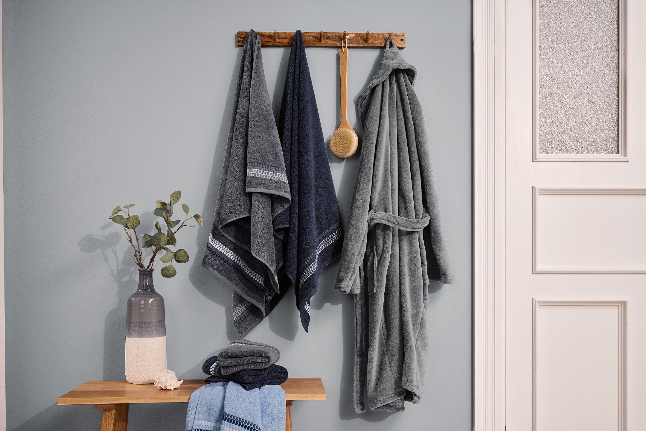 Grauer und blauer Bademantel und Handtücher hängen an einer Holzgarderobe, mit einer Vase und gefalteten Handtüchern auf einer Bank.