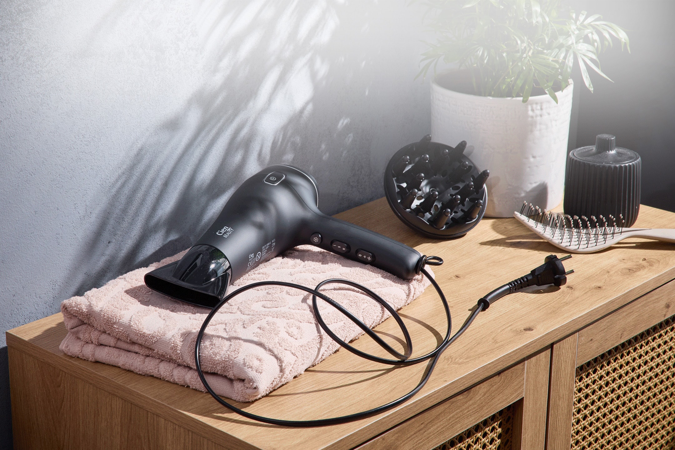 Asciugacapelli nero con diffusore, asciugamano rosa, spazzola e pianta su una cassettiera in legno.