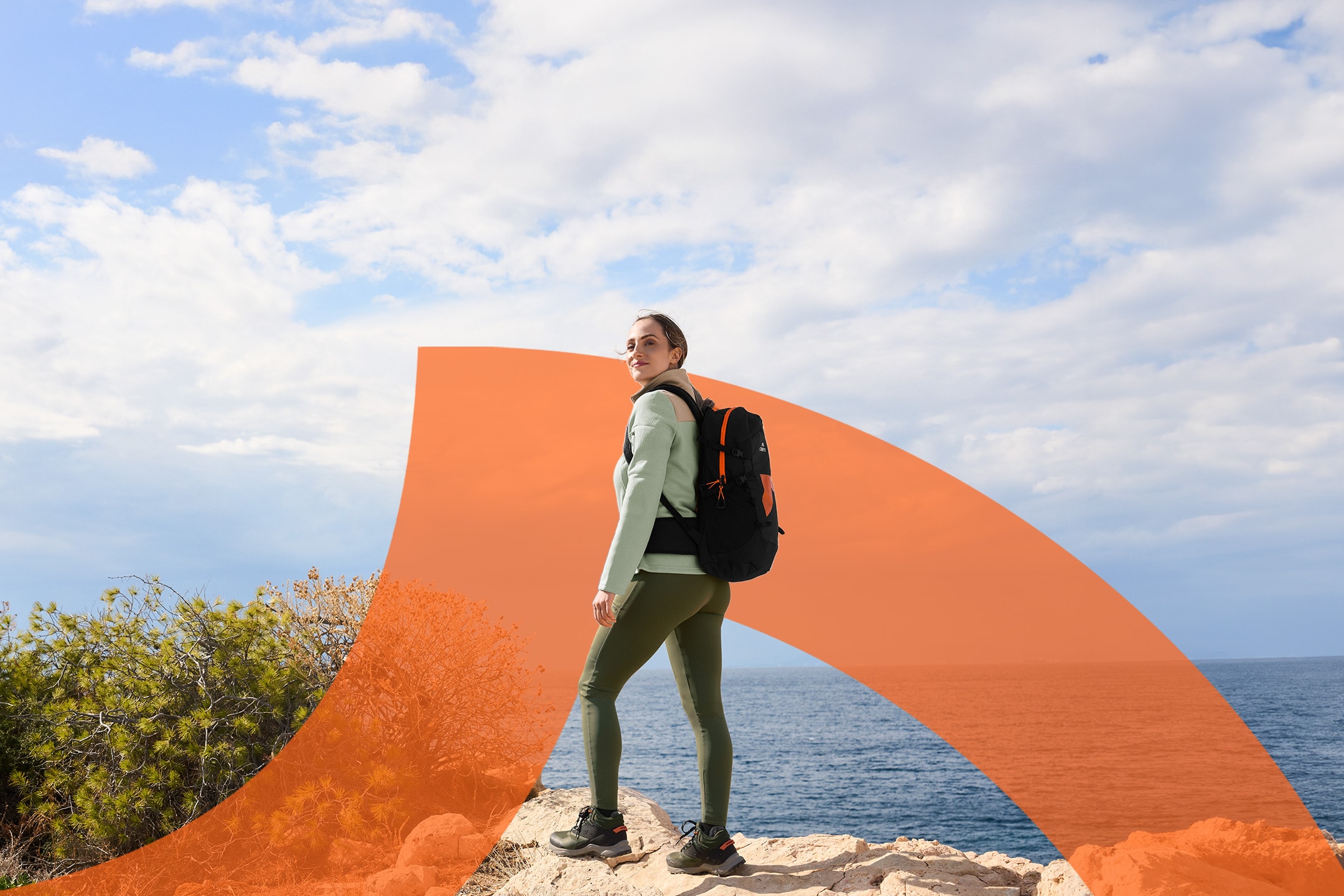 Frau in Wanderkleidung mit Rucksack, über die Schulter blickend, am Meer.