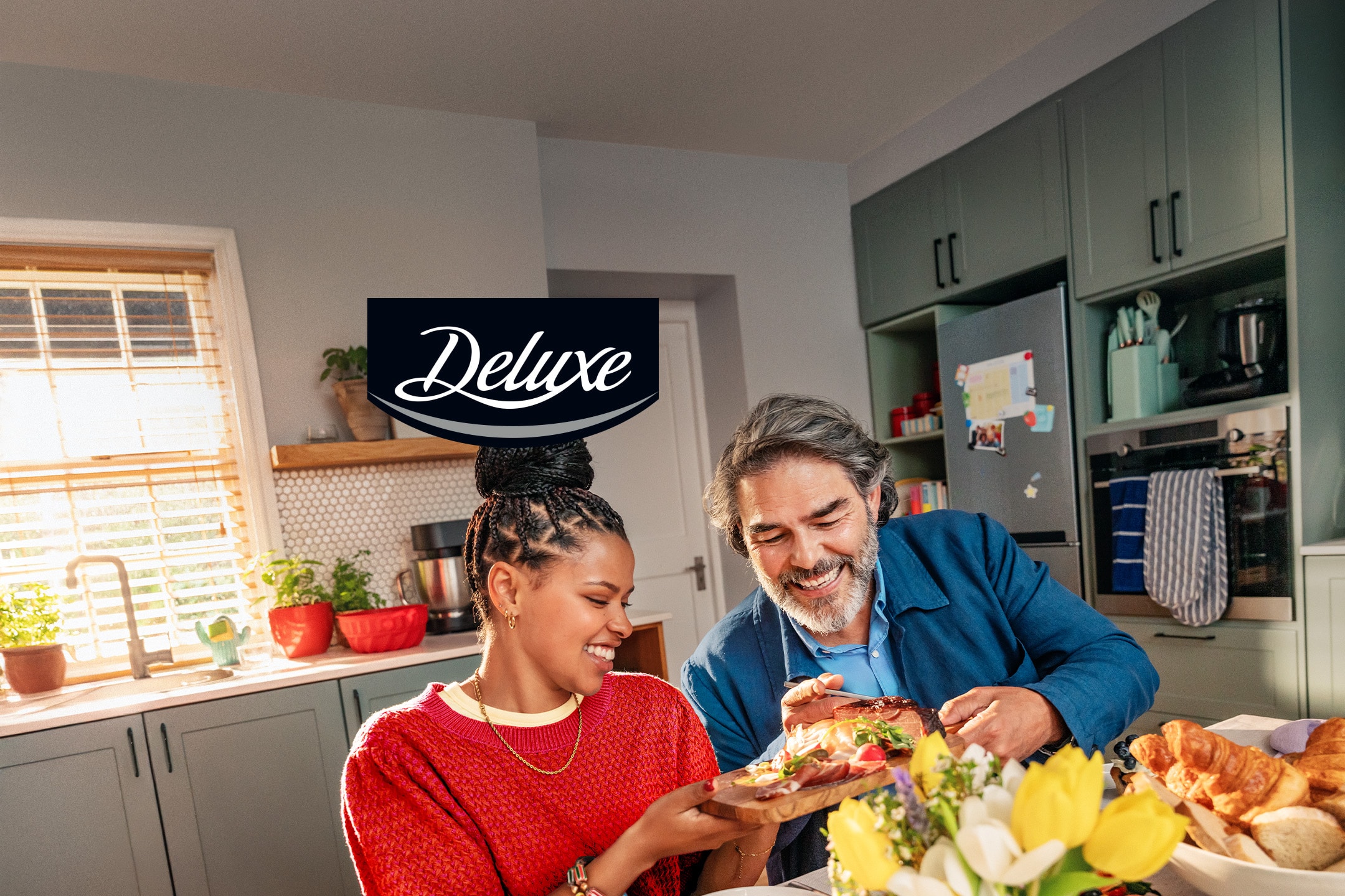 Un couple souriant partage un plateau de charcuterie et de fromages dans une cuisine lumineuse.