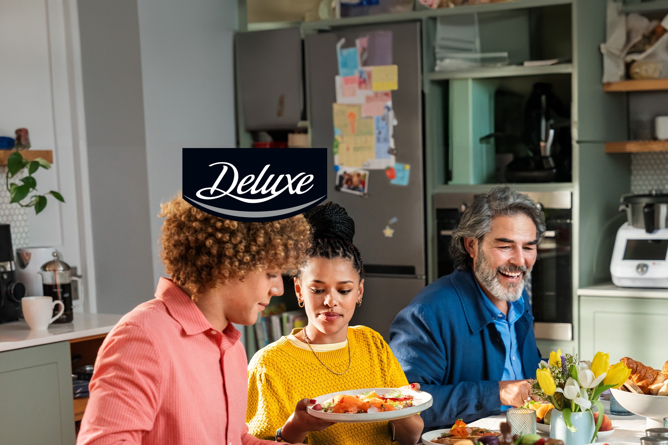 Drei Personen am Tisch mit einem reichhaltigen Frühstück, darunter Croissants und Blumen.