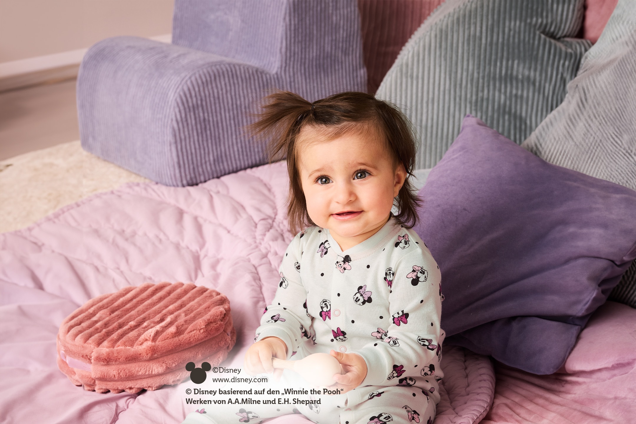 Baby in Minnie-Maus-Schlafanzug, sitzt auf einer rosa Decke mit Kissen.