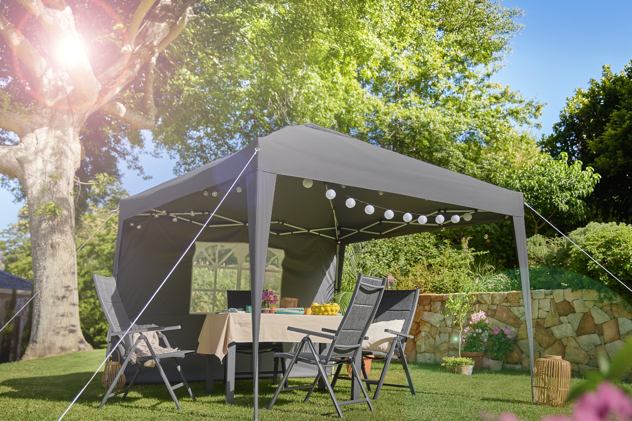 Graues Gartenpavillon mit Tisch und Stühlen, dekoriert mit Lichterketten, in einem sonnigen Garten.