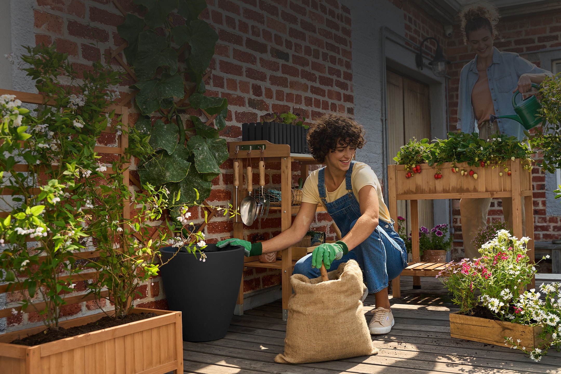 Due donne fanno giardinaggio su un balcone, con fioriere in legno e attrezzi da giardinaggio.