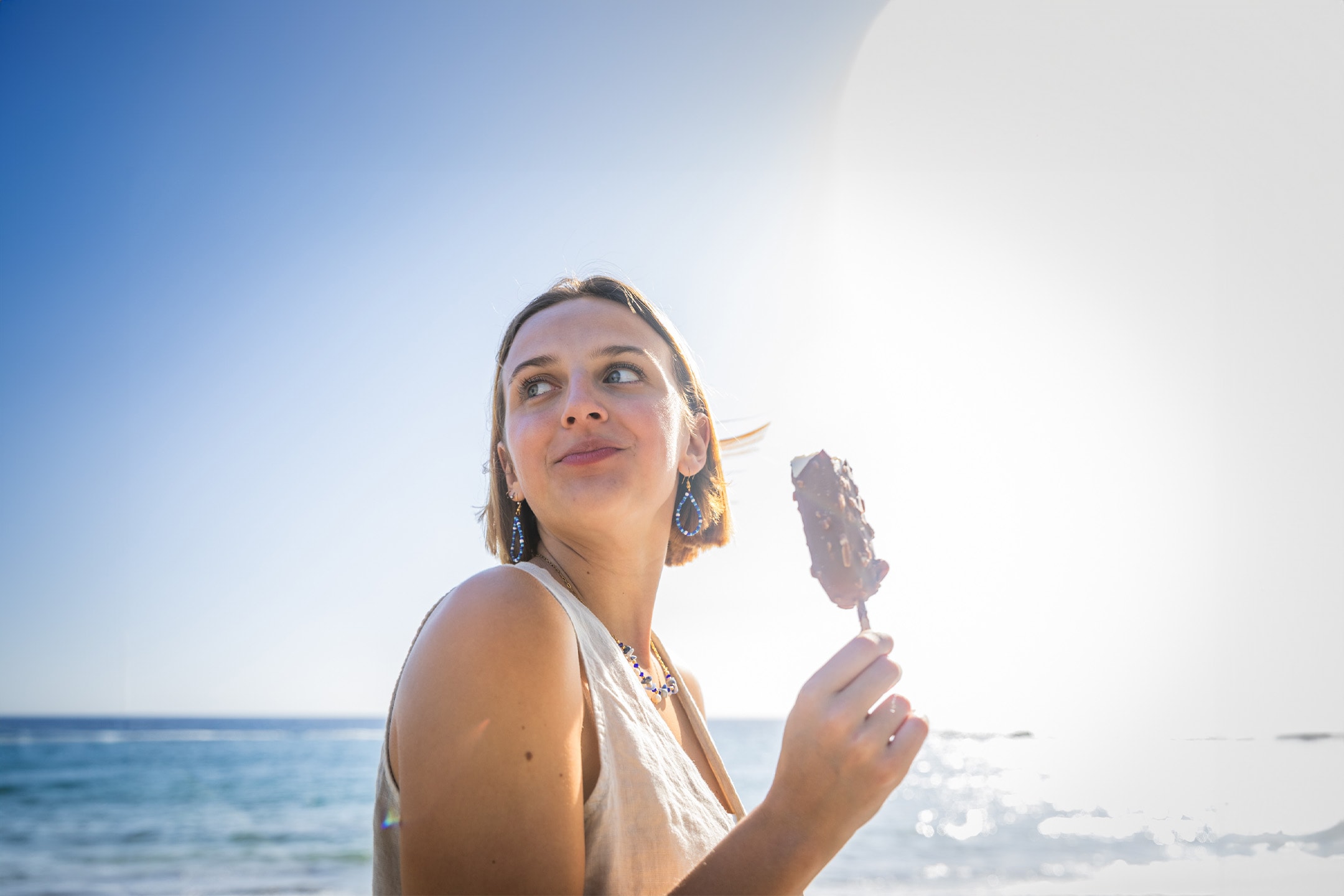 Frau am Strand mit einem Eis am Stiel, blickt in die Ferne.