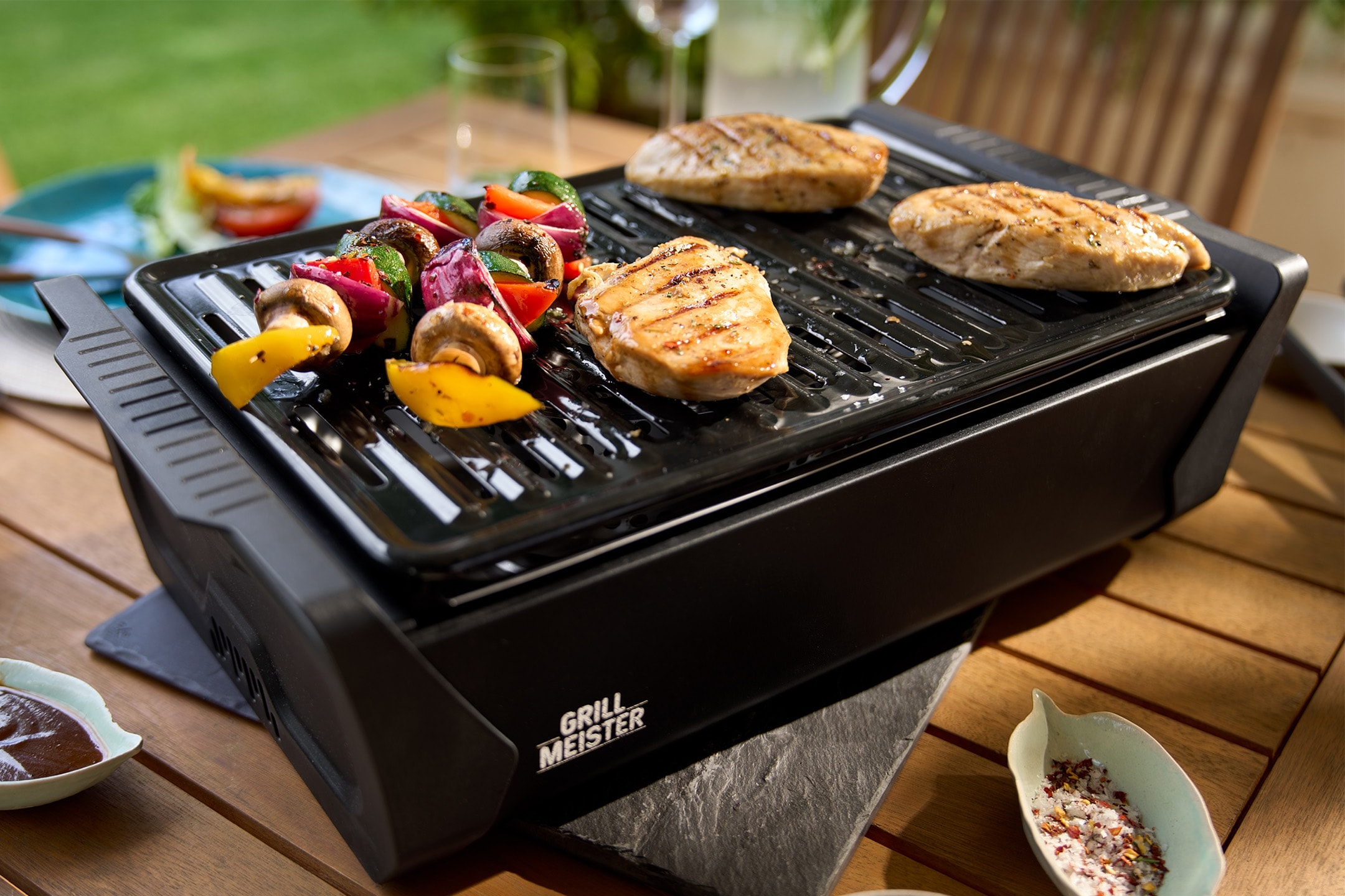 Griglia elettrica con pollo e spiedini di verdure su un tavolo di legno all'aperto.