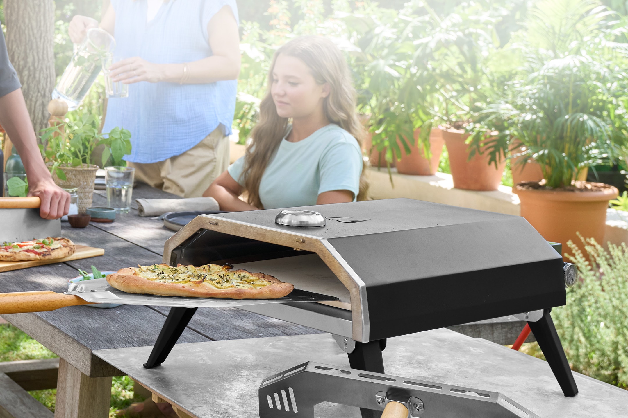 Outdoor-Pizzaofen mit backender Pizza auf einem Holztisch mit Personen.