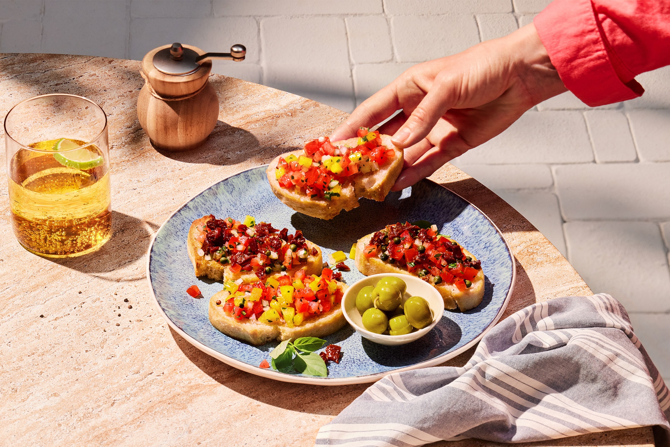 Eine Hand greift nach Bruschetta mit gewürfelten Tomaten und Paprika, neben einem Getränk und Oliven.