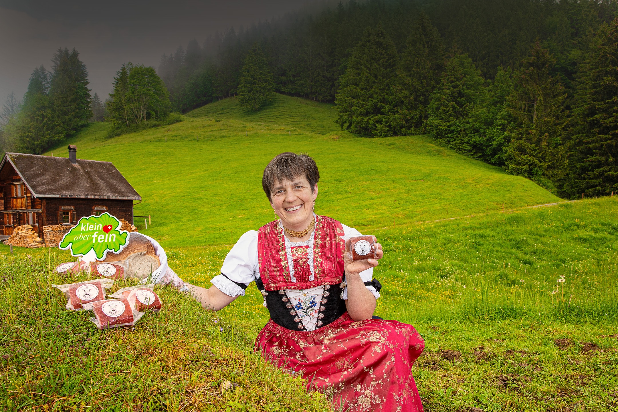 Frau in Tracht präsentiert Fleischprodukte und Brot vor Almhütte und Wald, mit Schild 'klein aber fein'.