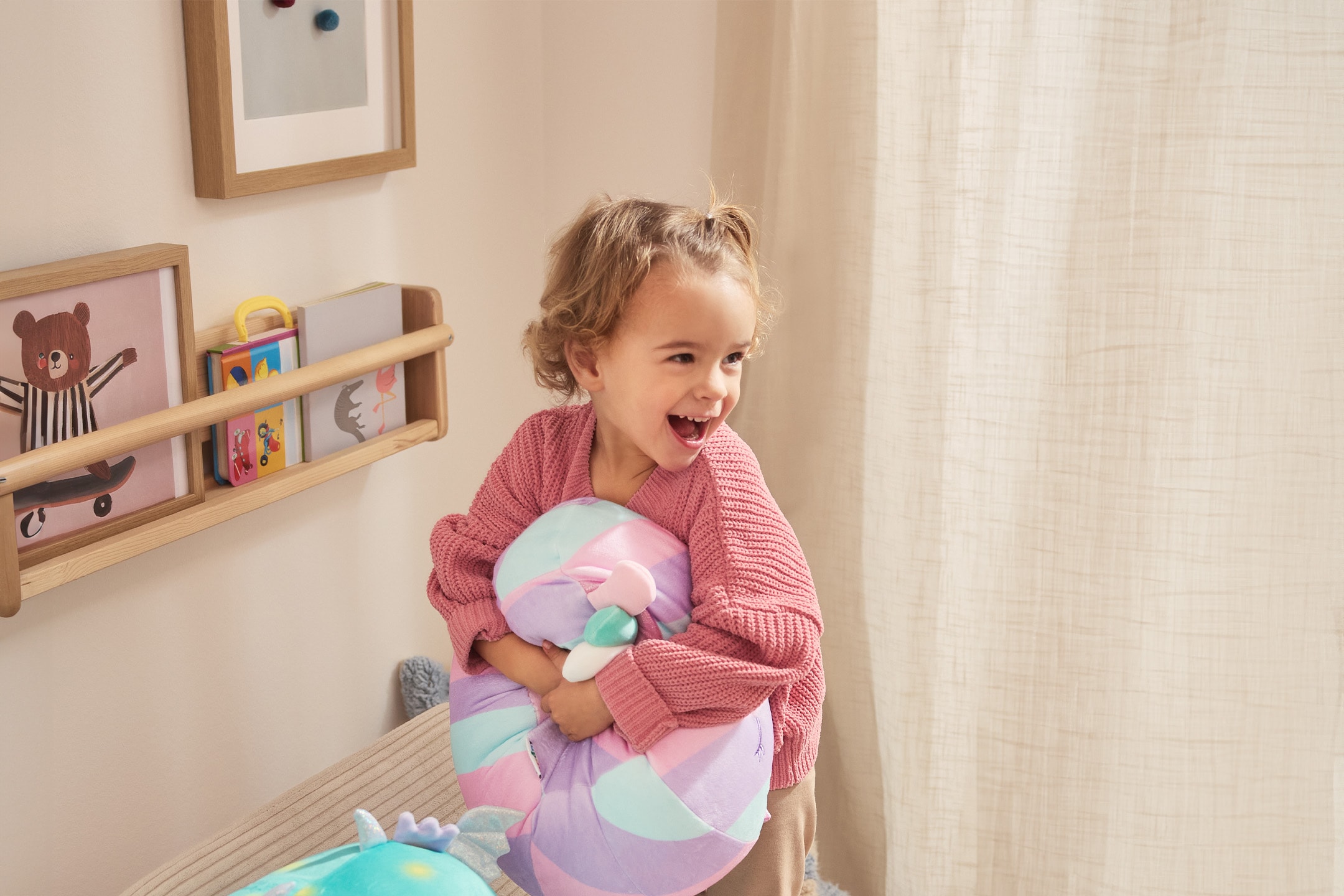Petite fille joyeuse en pull rose serrant un coussin coloré dans une chambre avec étagères.