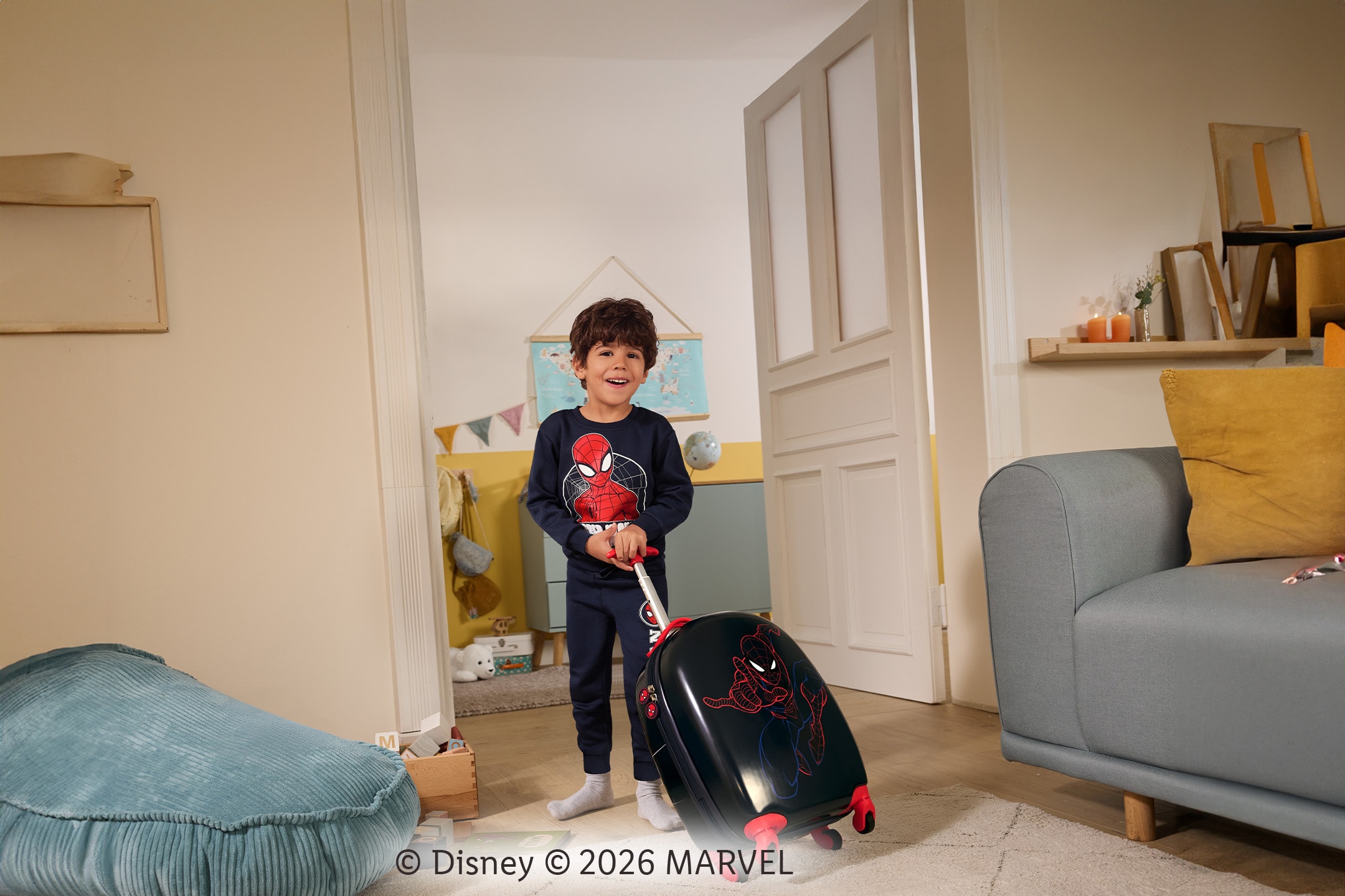 Ein lächelndes Kind im Spiderman-Schlafanzug zieht einen Spiderman-Koffer in einem Wohnzimmer.