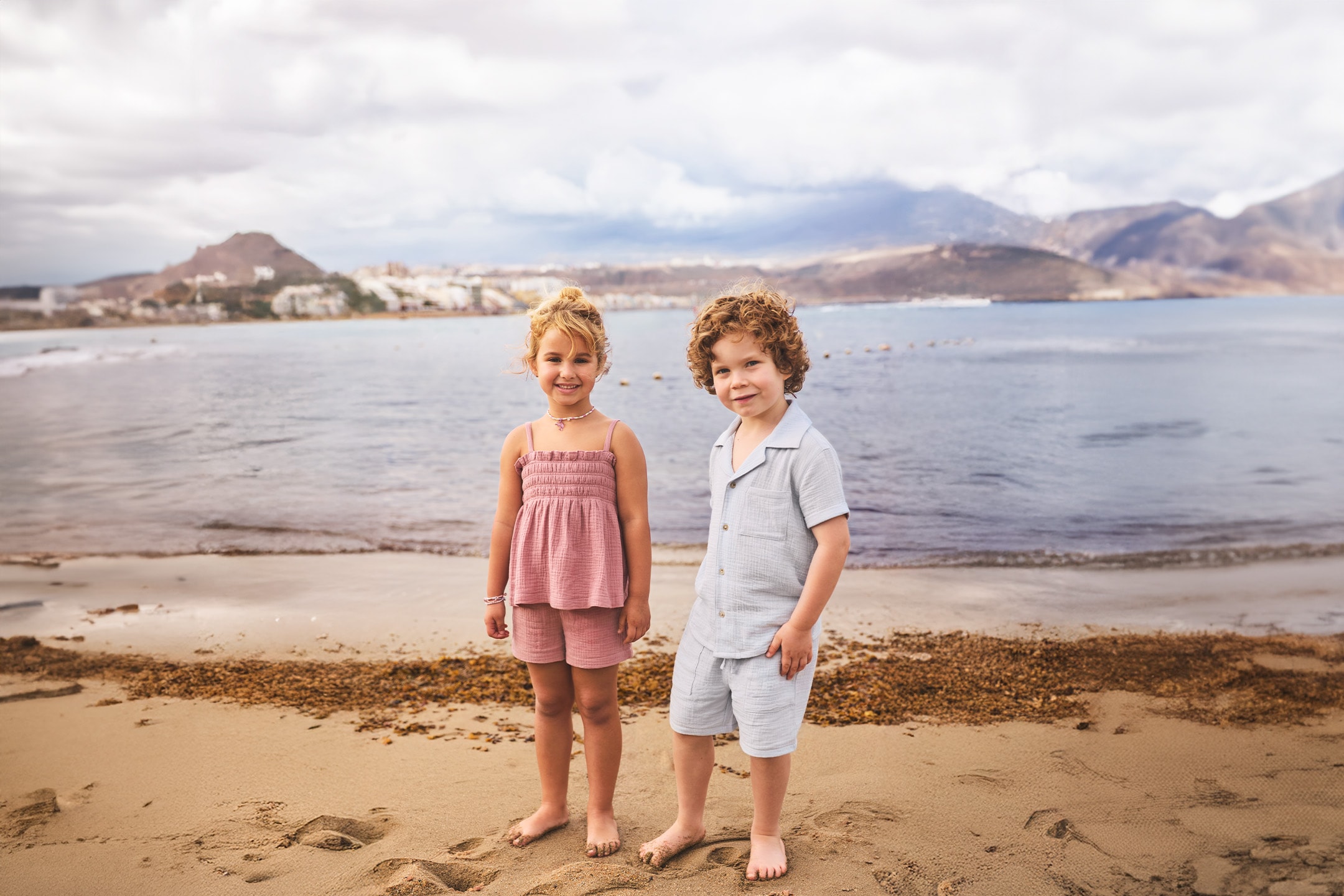 Due bambini in abiti estivi sulla spiaggia con il mare e le montagne sullo sfondo.
