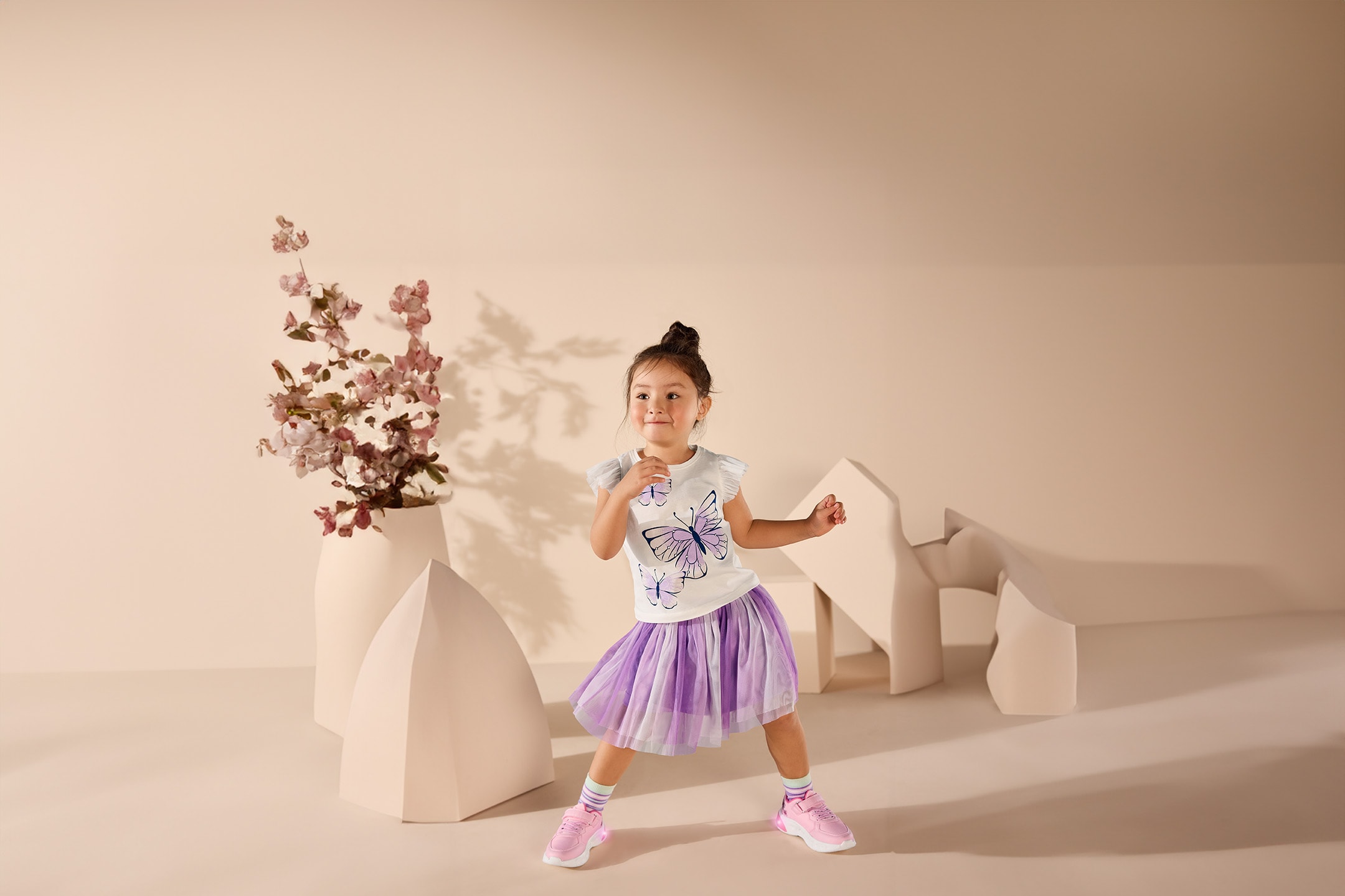 Kleines Mädchen in weißem T-Shirt mit Schmetterlingen und lila Tüllrock, posiert in einem Raum mit beigen Dekorationen.