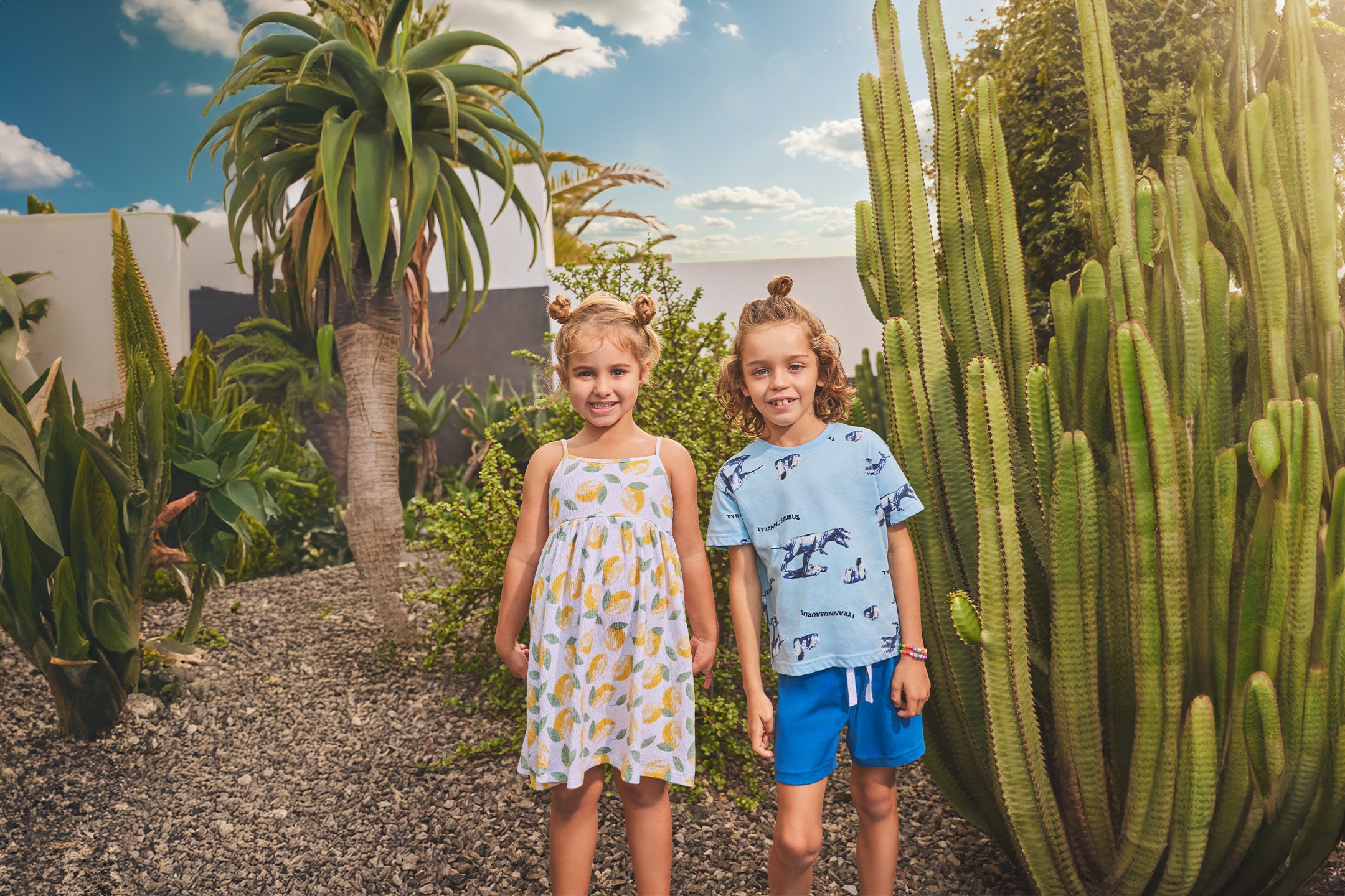 Due bambini sorridenti, una bambina in vestito a limoni e un bambino in maglietta dinosauri e pantaloncini blu, in un giardino di cactus.