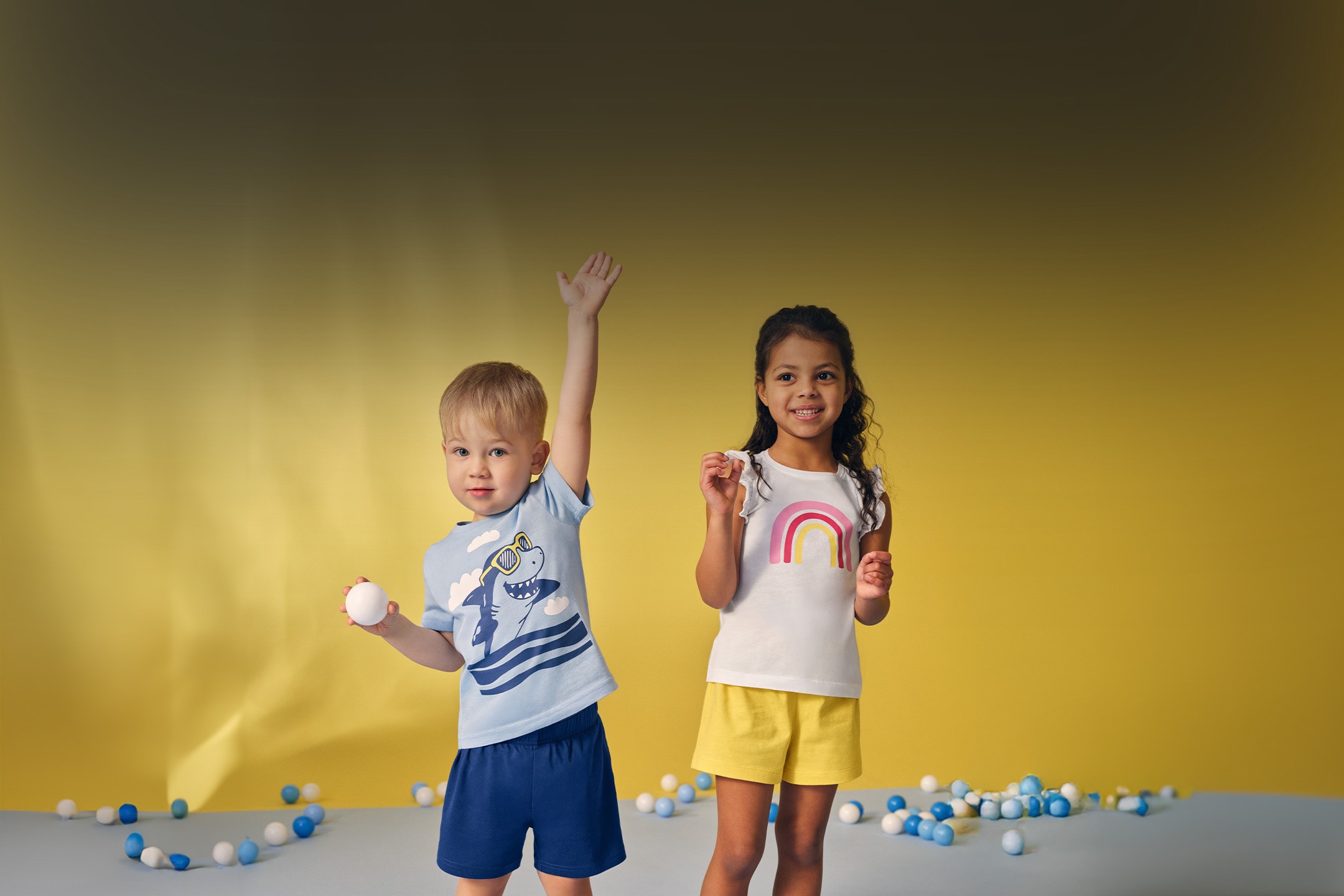 Zwei lächelnde Kinder tragen Sommer-T-Shirts und -Shorts, umgeben von Bällen.