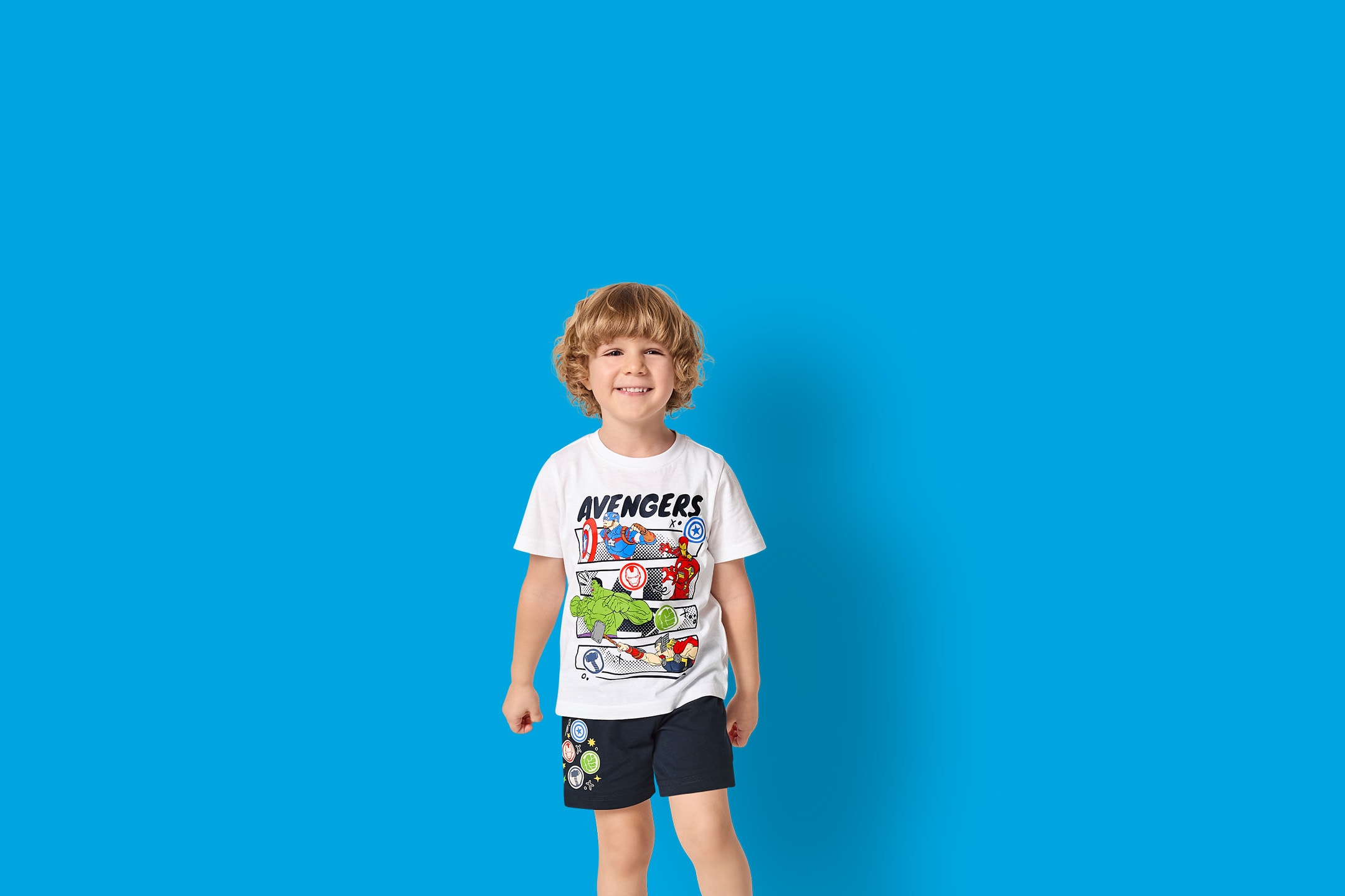 Bambino in maglietta bianca con motivo di supereroi e pantaloncini blu su sfondo blu.