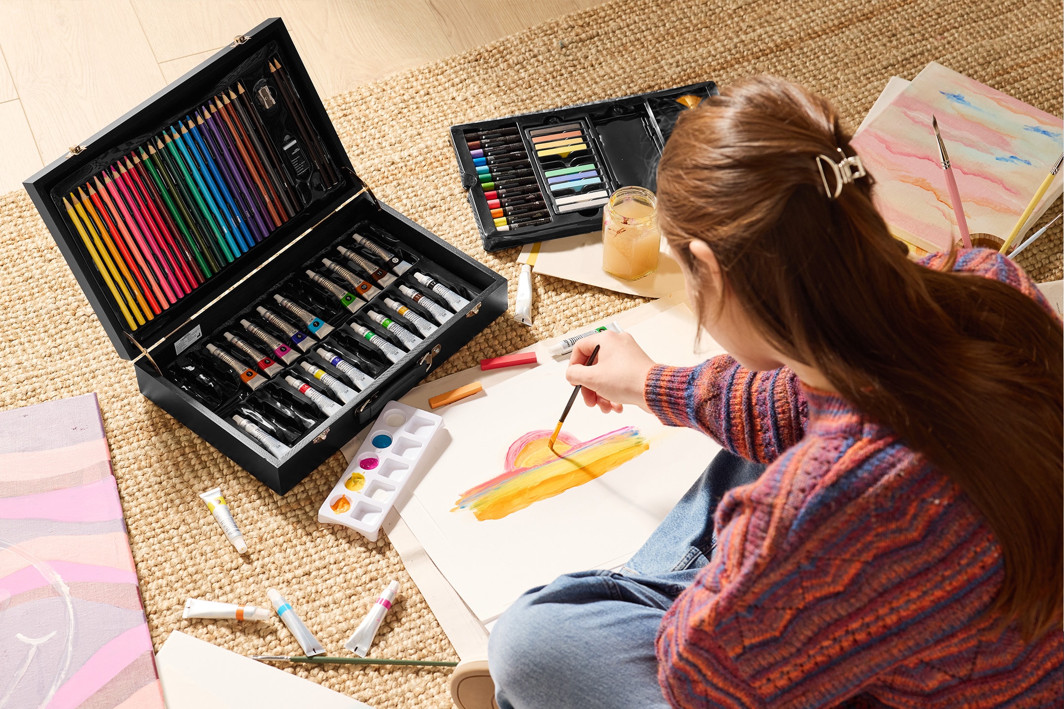 Eine Person malt mit einem Kunstset, das Buntstifte, Farbtuben und Pastellkreiden enthält.