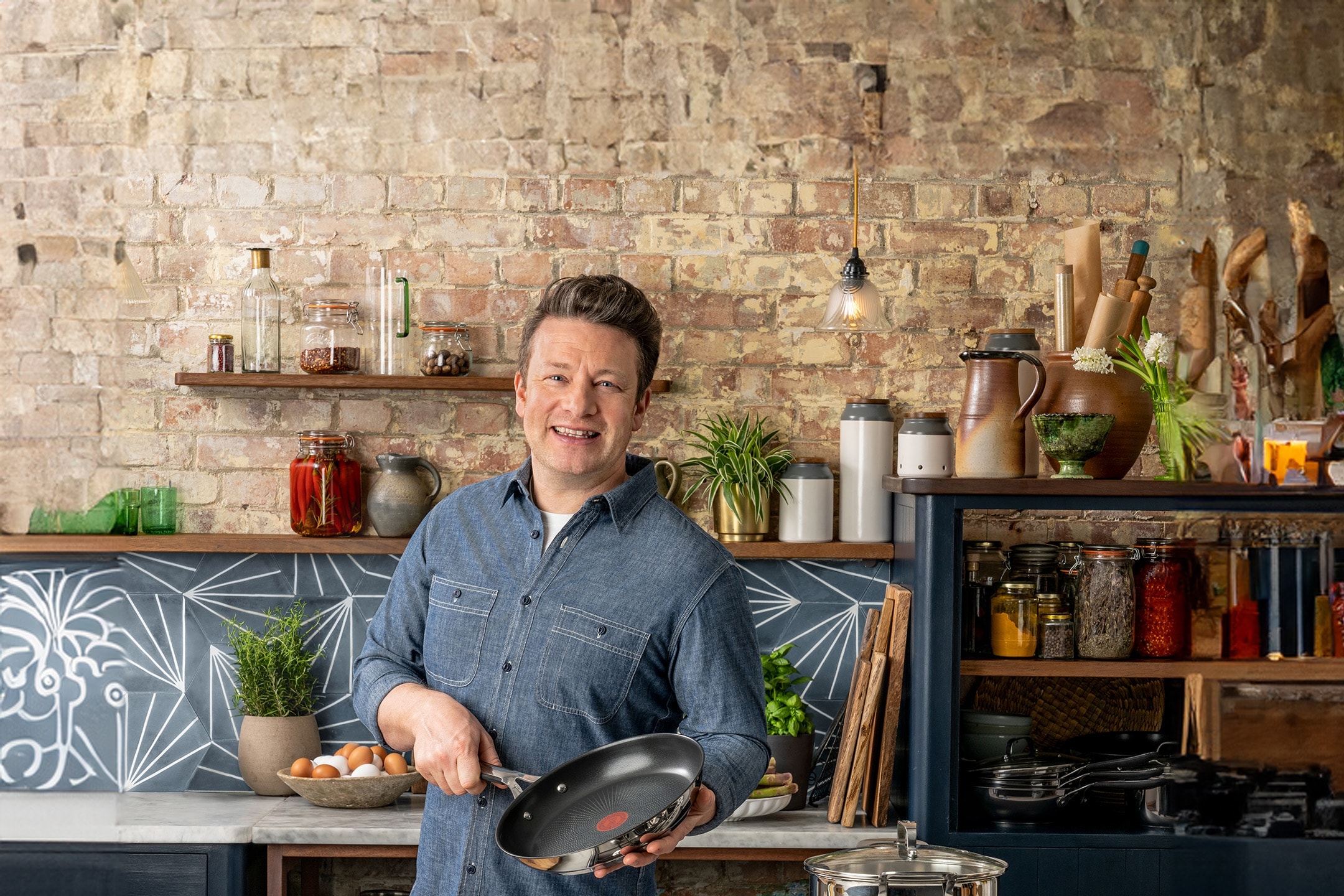 Ein lächelnder Mann hält eine Pfanne in einer rustikalen Küche mit Regalen voller Zutaten.