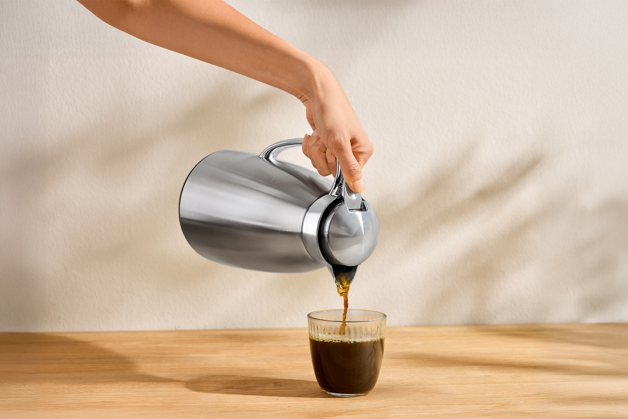 Une main verse du café d'une verseuse isotherme en acier inoxydable dans un verre sur une table en bois.