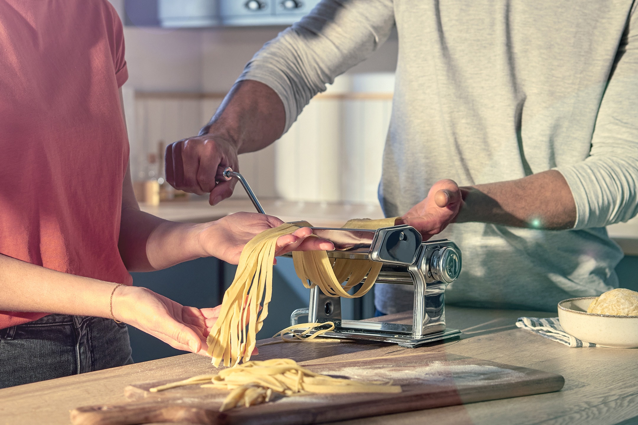 Zwei Personen verwenden eine Nudelmaschine, um frische Tagliatelle auf einem Holzschneidebrett zuzubereiten.
