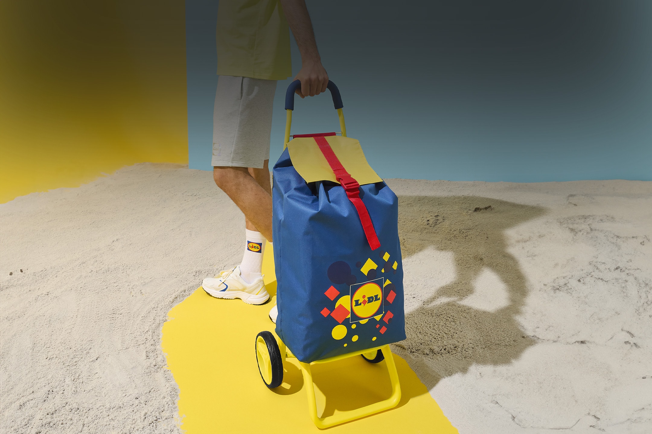 Ein blauer und gelber Einkaufstrolley mit geometrischem Muster, von einer Person am Strand gehalten.
