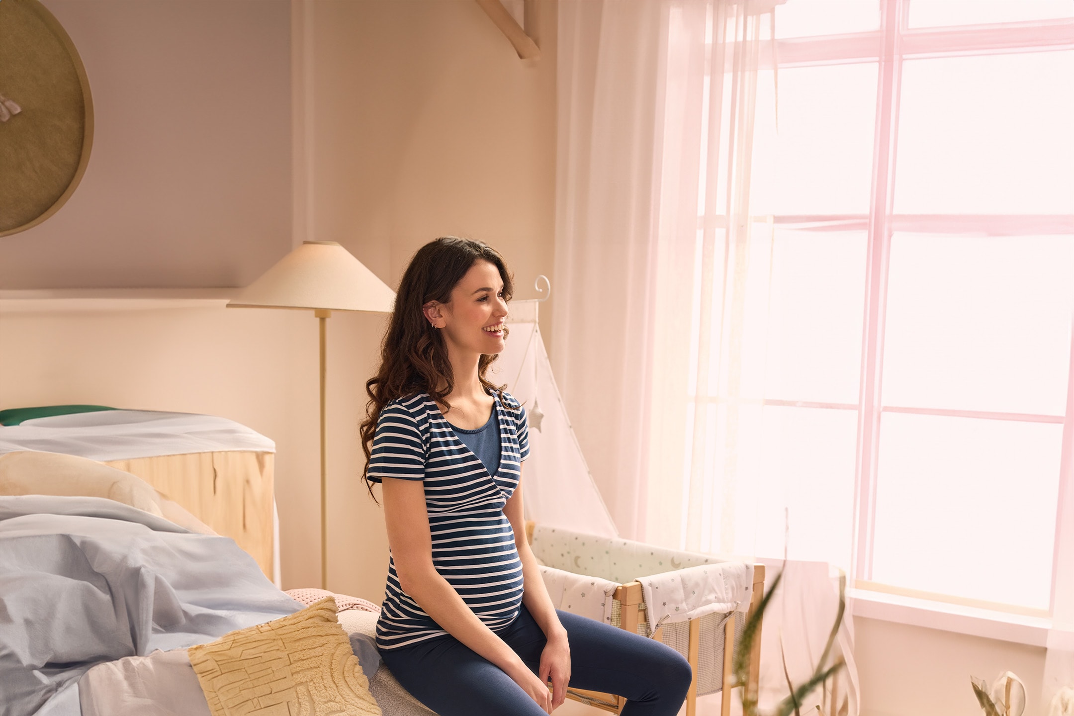 Lächelnde schwangere Frau in gestreiftem T-Shirt und Leggings, auf dem Bett in einem hellen Schlafzimmer sitzend.