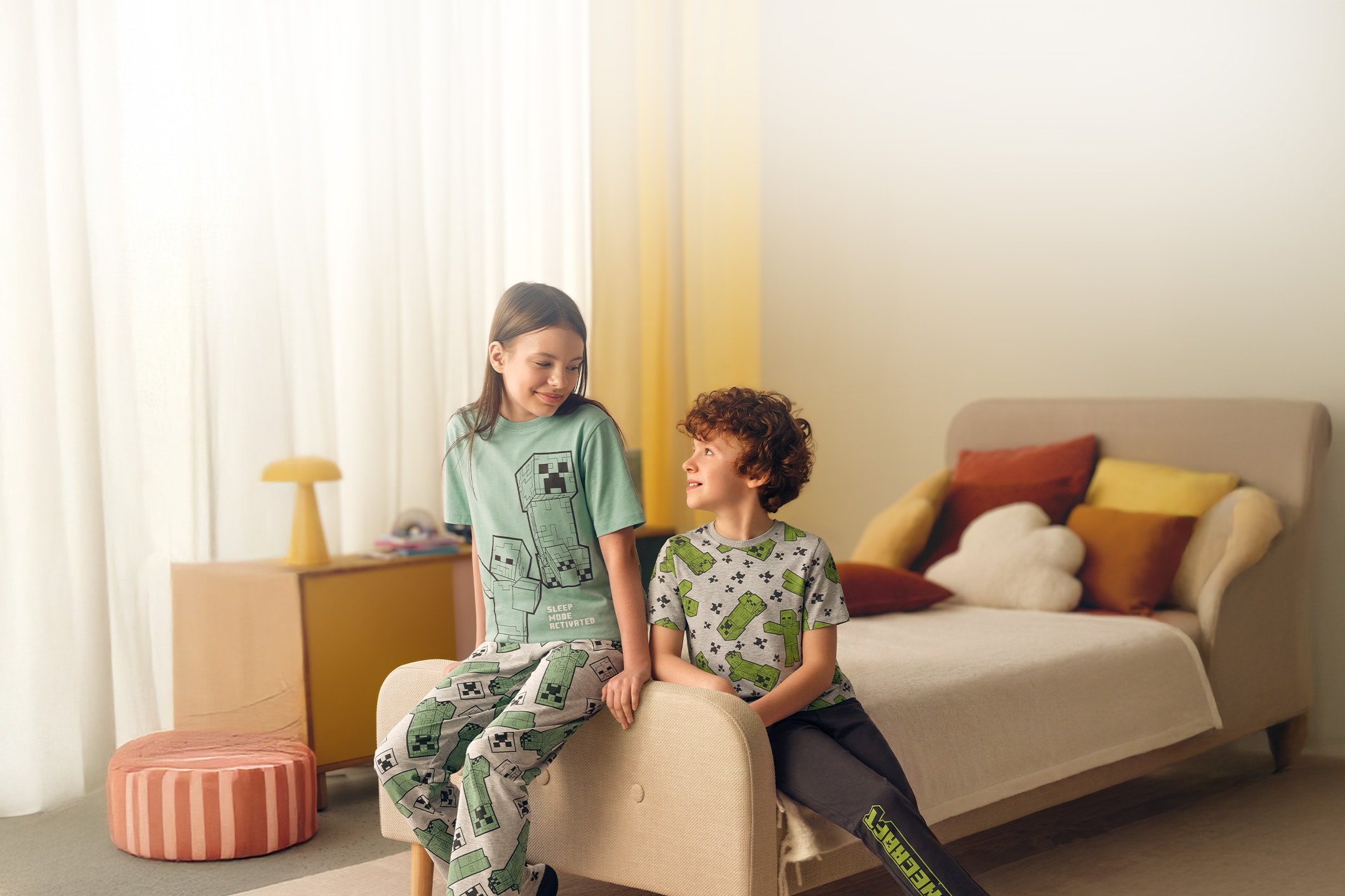 Zwei Kinder in Pyjamas mit Videospiel-Motiven sitzen auf einem Bett in einem hellen Schlafzimmer.