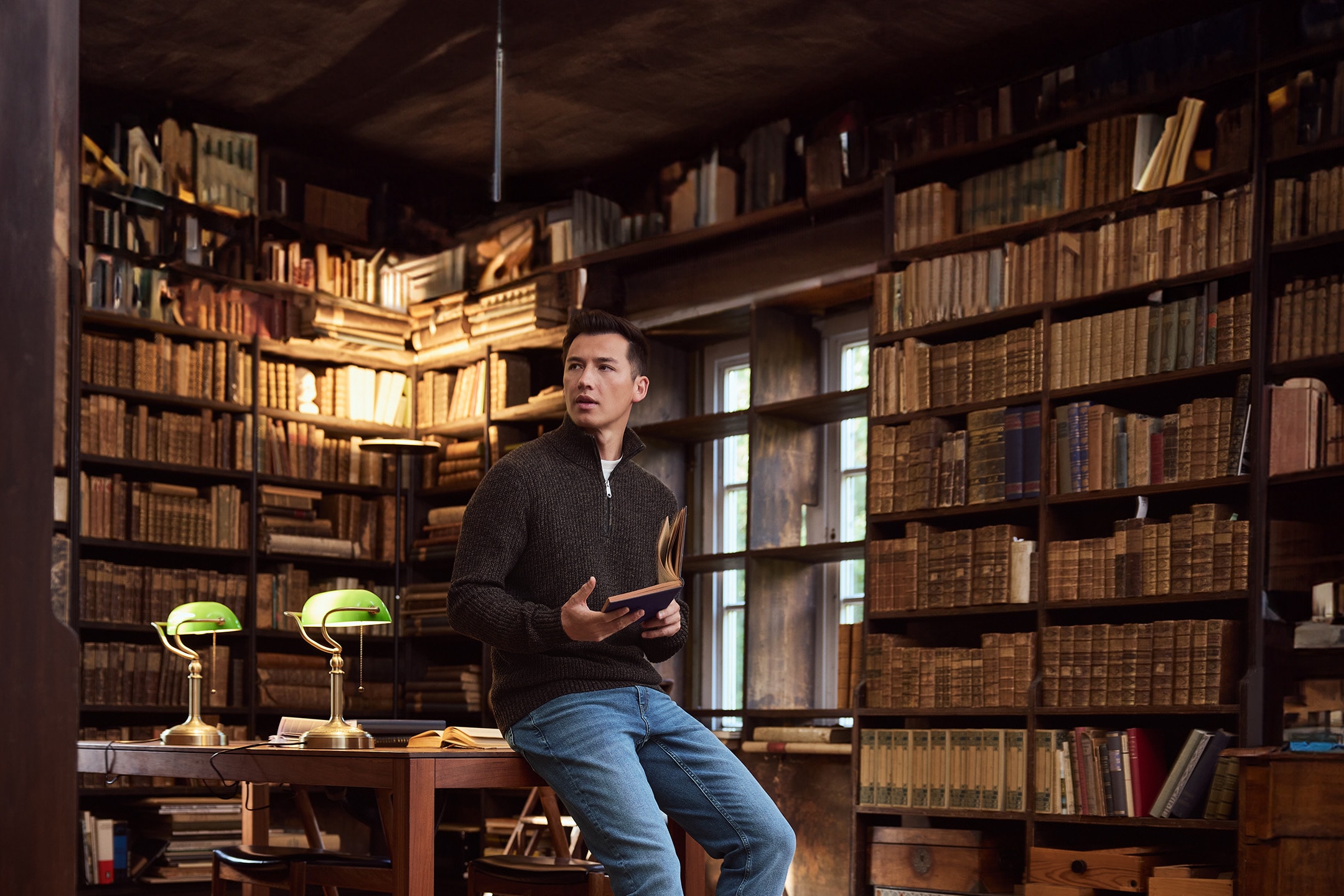 Mann in Strickpullover und Jeans liest in einer Bibliothek mit vielen Büchern.